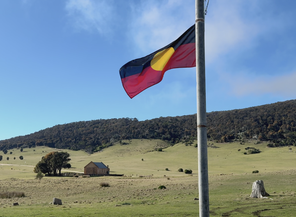 A photo of the Aboriginal Flag flying in the foreground of the Wybalenna chapel on Flinders Island