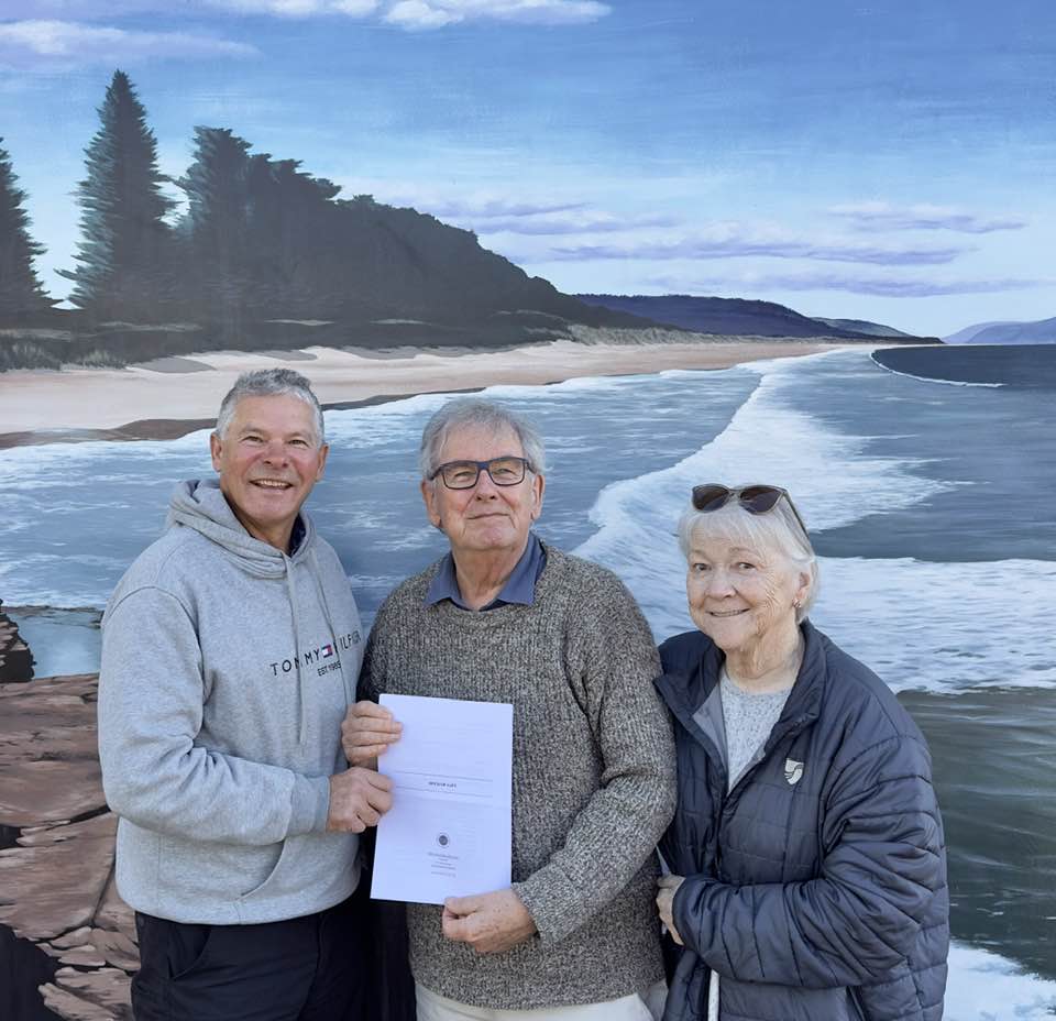 Three people stand in front of a mural of the sea. The man on the left and the man in the middle are holding a document that transfers a title of land to the Aboriginal Community. The third person is a woman standing on the right next to them.